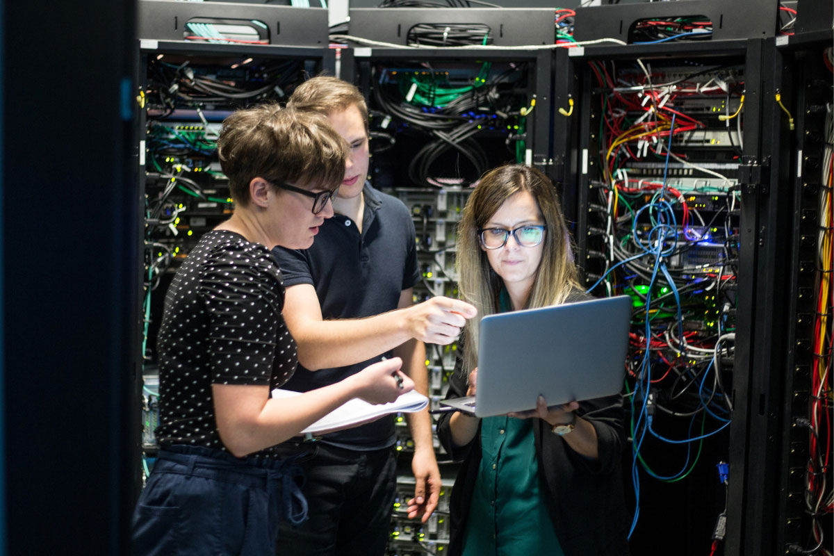 A team of three IT professionals collaborating on a laptop inside a server room, troubleshooting a complex network issue