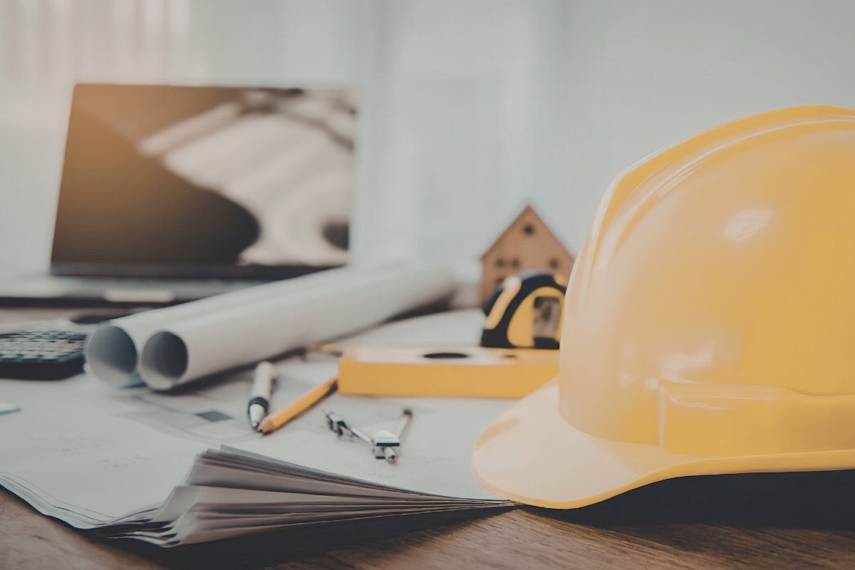 Yellow hard hat on a desk beside blueprints, measuring tools, and a laptop, representing infrastructure and construction planning.
