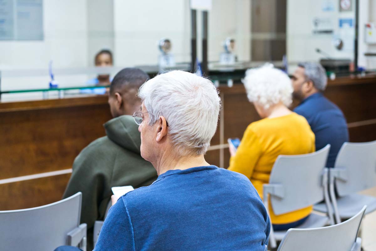Patients seated in a healthcare waiting room, some using mobile phones while waiting for service.